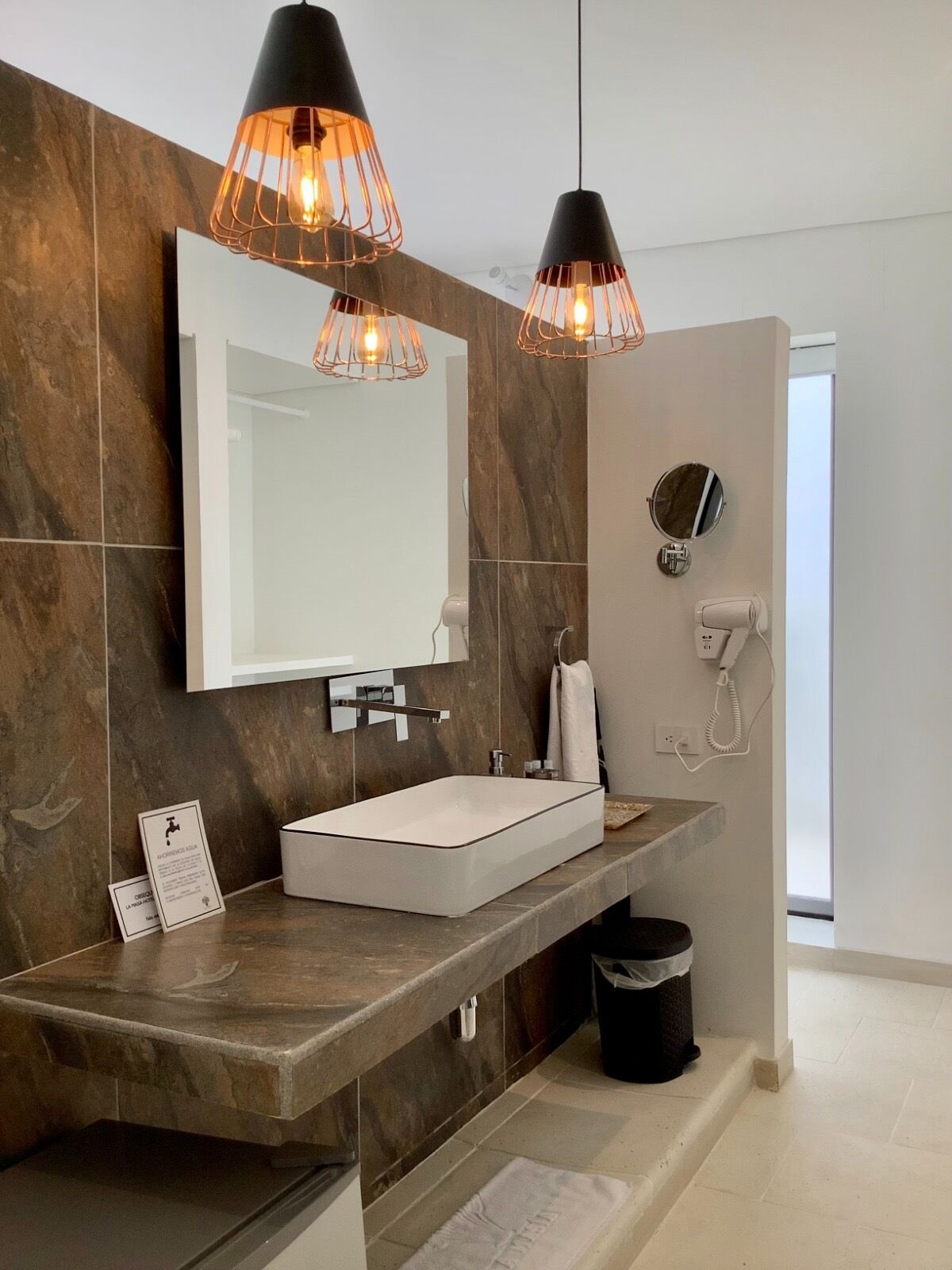 Un baño moderno con un espejo grande, un lavamanos rectangular y dos lámparas colgantes con bombillas a la vista.