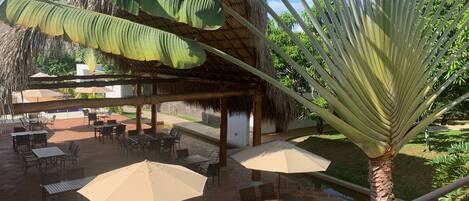 An outdoor patio area with tables and chairs, a thatched roof structure, and a palm tree.
