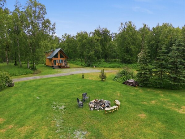 A cabin with a fire pit and chairs in a grassy yard, surrounded by trees.