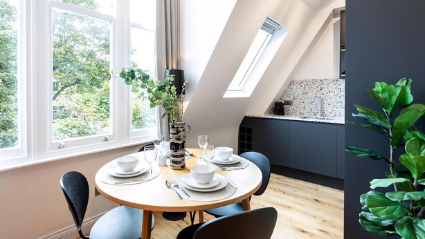 A dining area with a round table set for four, large windows, a skylight, and a view of greenery outside. - Coleherne Road Residence X (London)