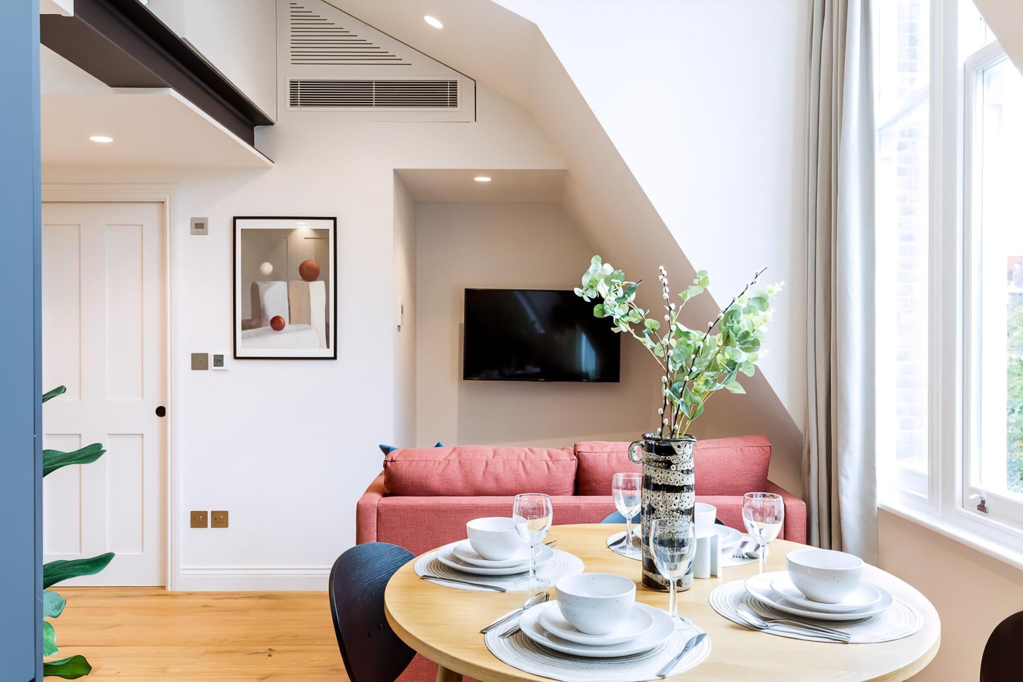 A dining area with a round table set for four, a red sofa, a TV mounted on the wall, and a window with curtains.