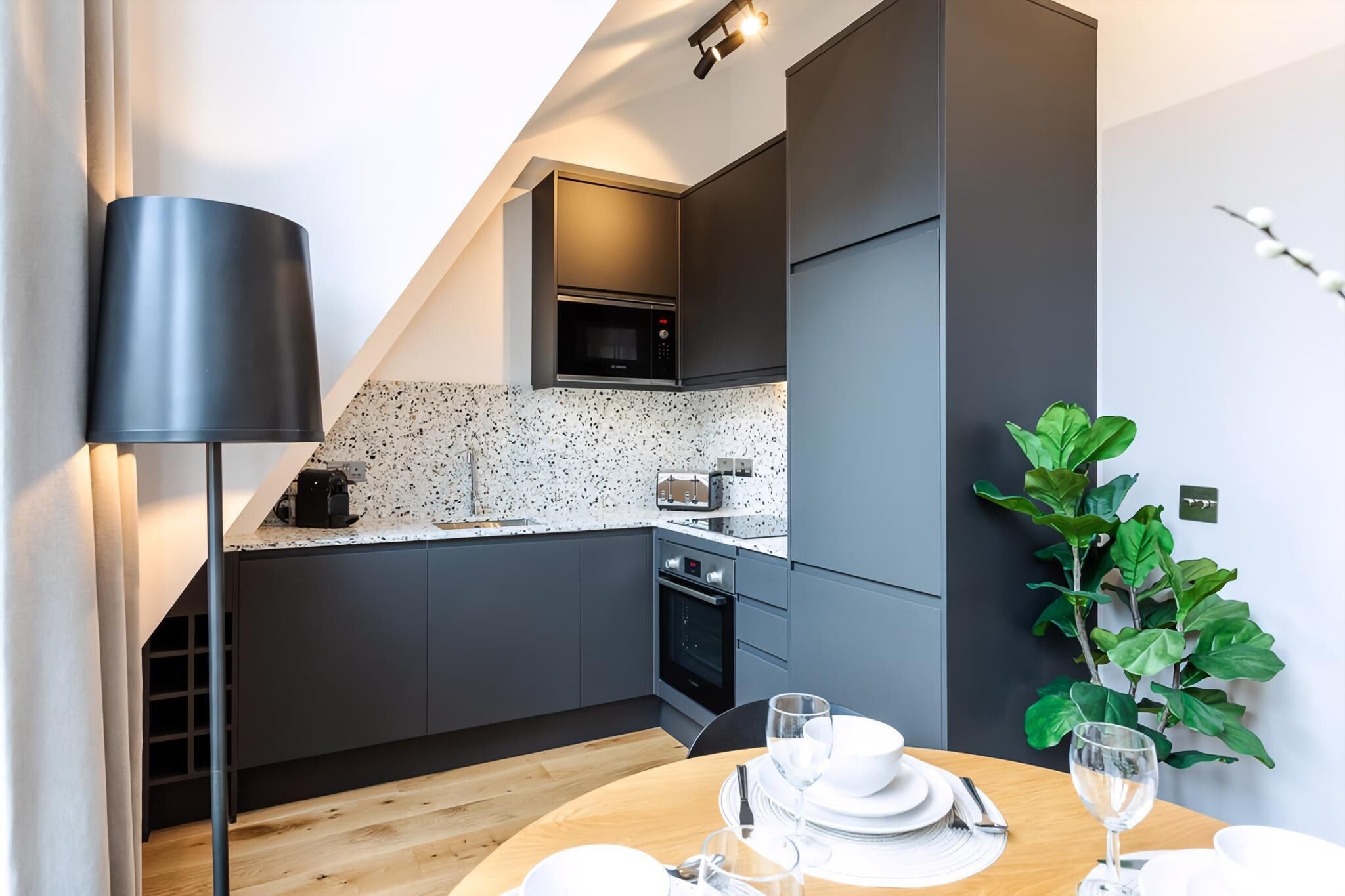 A modern kitchen with dark grey cabinets, a microwave, a toaster, and a wooden dining table set with plates, glasses, and cutlery.