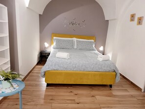 A bedroom with a bed, a nightstand, a lamp, a plant, and a wall with a floral design. - Centro Città My House (Catania)