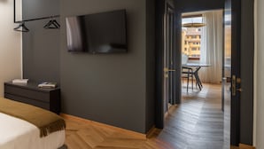A modern hotel room with a large flat-screen TV mounted on the wall, a wooden herringbone floor, a bed with a white comforter and brown throw, and a view of the city through an open door. - Oro1 Florence Luxury Apartment by Mmega (Florence)