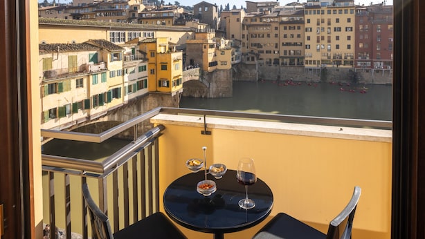 A balcony with a table, two chairs, and a view of a river with boats. - Oro1 Florence Luxury Apartment by Mmega (Florence)