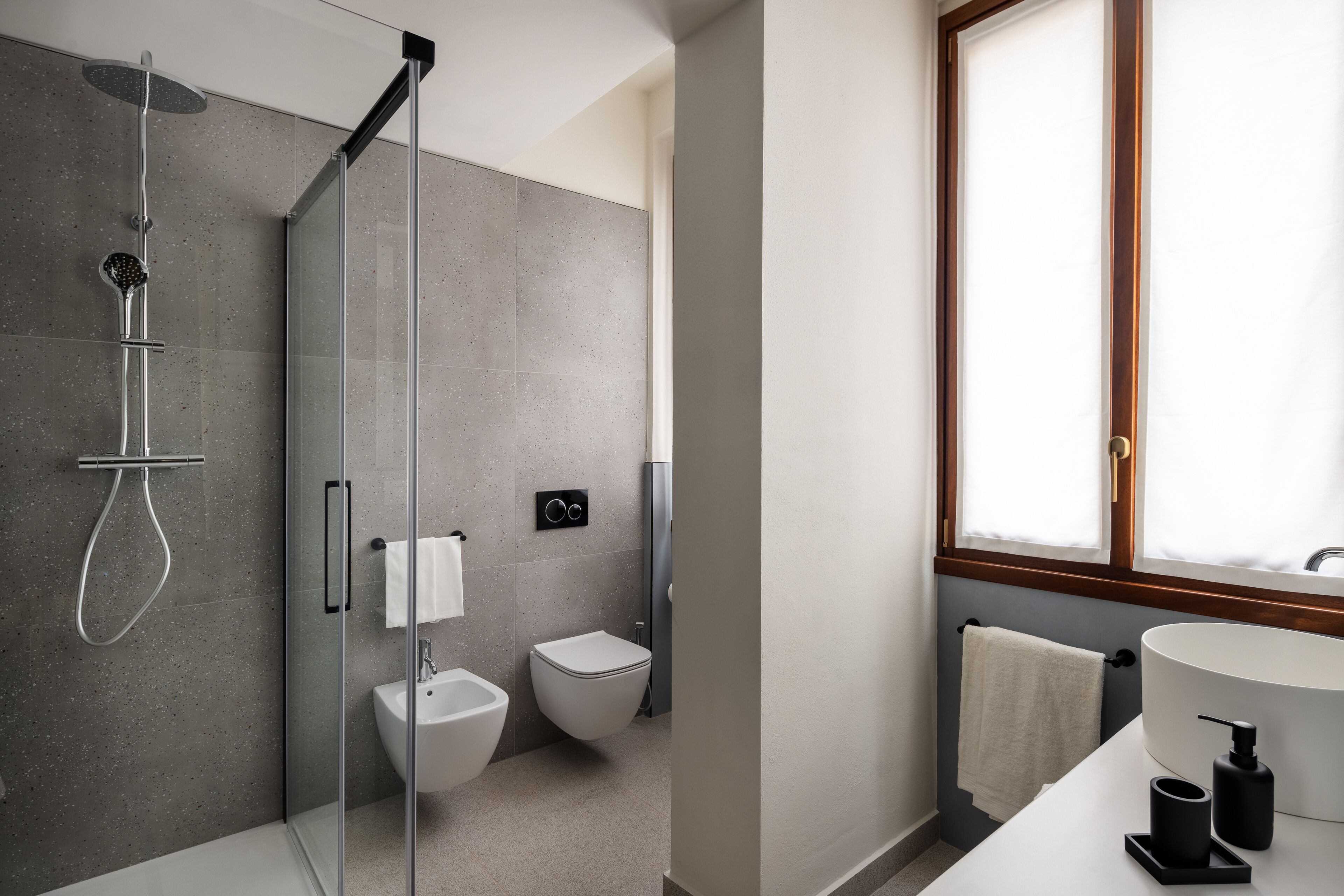 A modern bathroom with a glass-enclosed shower, a bidet, a toilet, and a sink with a window above.