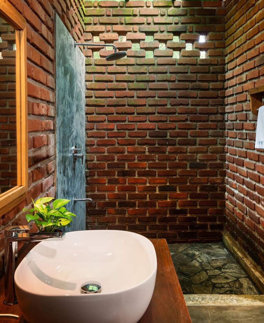 Een badkamer met een wit wastafel, een houten wastafelmeubel, een groene plant en een bakstenen muur.