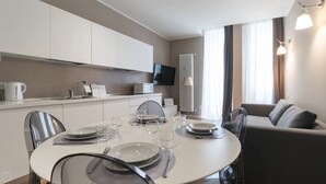 A modern dining area with a round table set for four, a flat-screen TV mounted above the counter, and a sofa in the background.