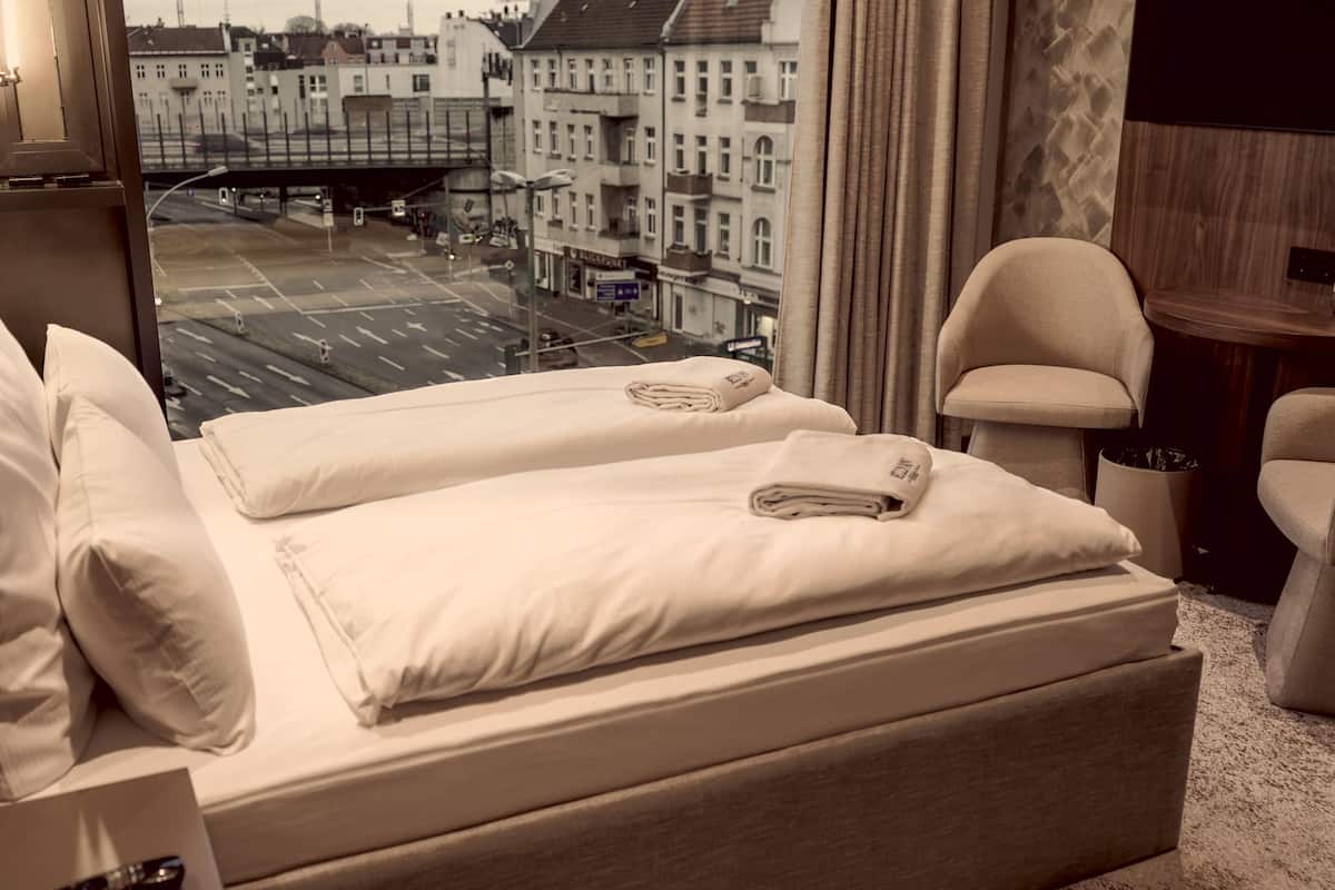 A hotel room with a bed, two pillows, a bedside table, a chair, and a view of a cityscape.