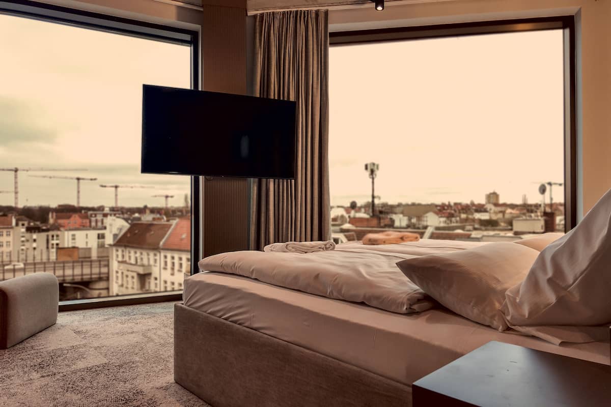 A bedroom with a large window, a flat-screen TV, a bed with beige bedding, and a view of a cityscape.