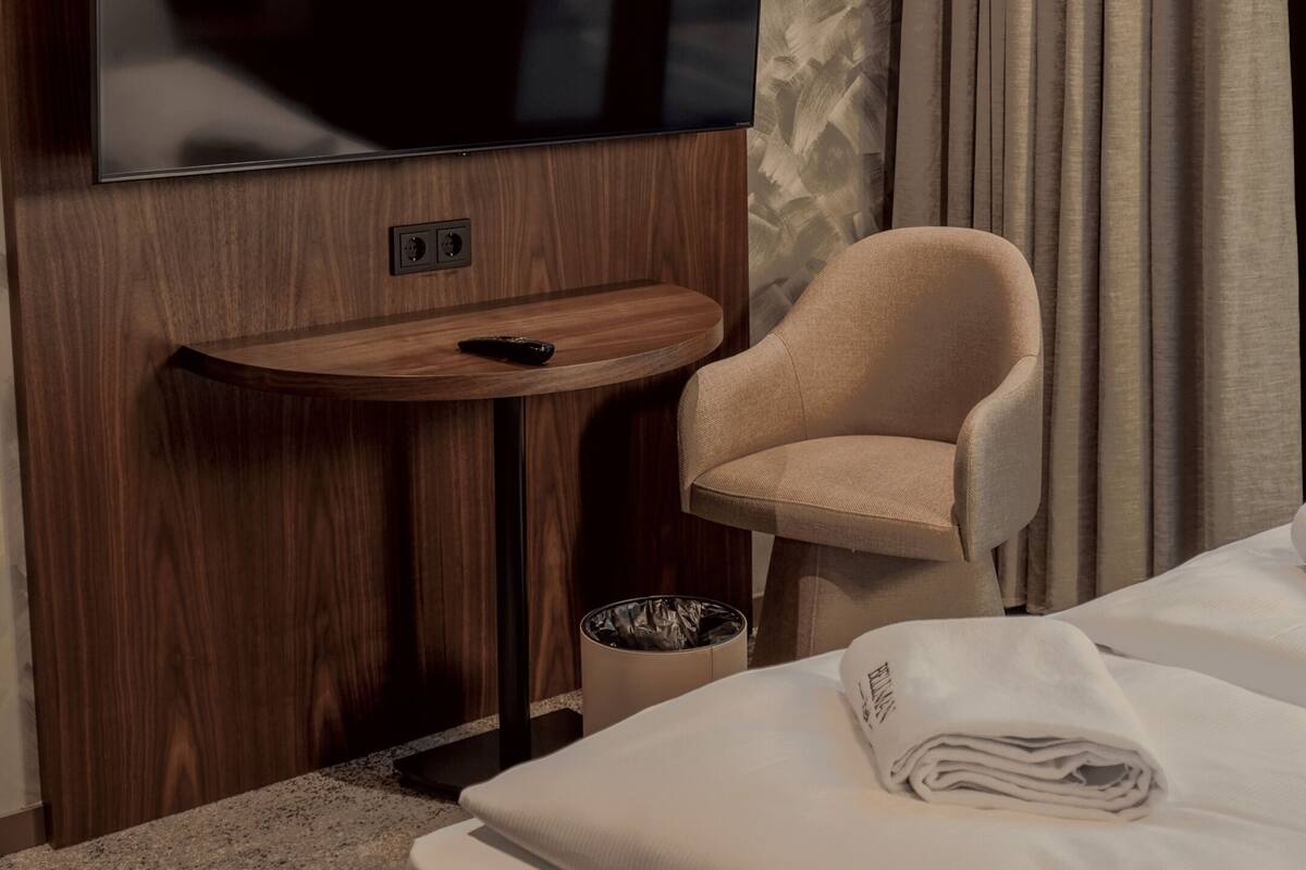 A hotel room with a flat-screen TV mounted on a wooden panel, a small wooden table with a remote control, a beige armchair, a trash bin, and a neatly made bed with a folded towel.