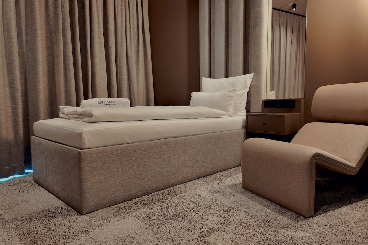A neatly made bed with a pillow labeled "BELLMAN Hotels," a bedside table with a lamp, and a chair in a hotel room.