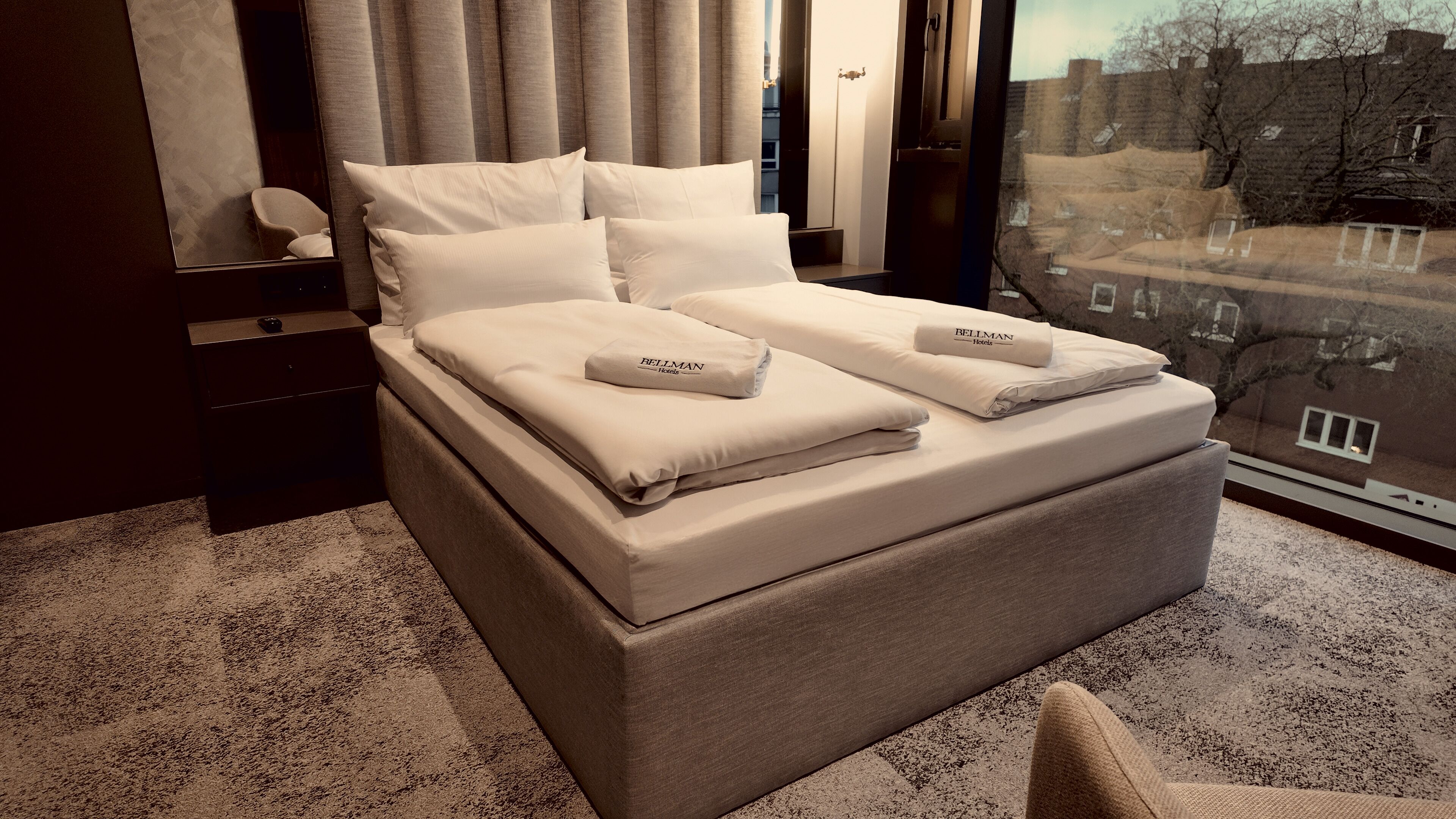 A double bed with white linens and pillows, a bedside table, a chair, and a large window with a view of buildings.
