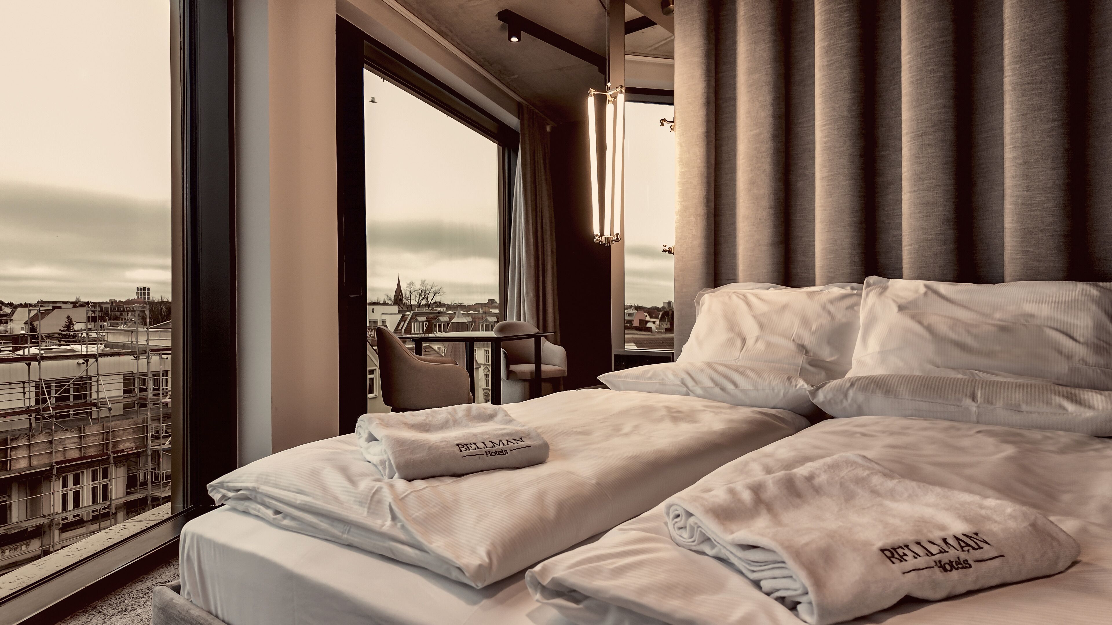 A hotel room with a large window offering a city view, a neatly made bed with pillows, and a table with chairs.