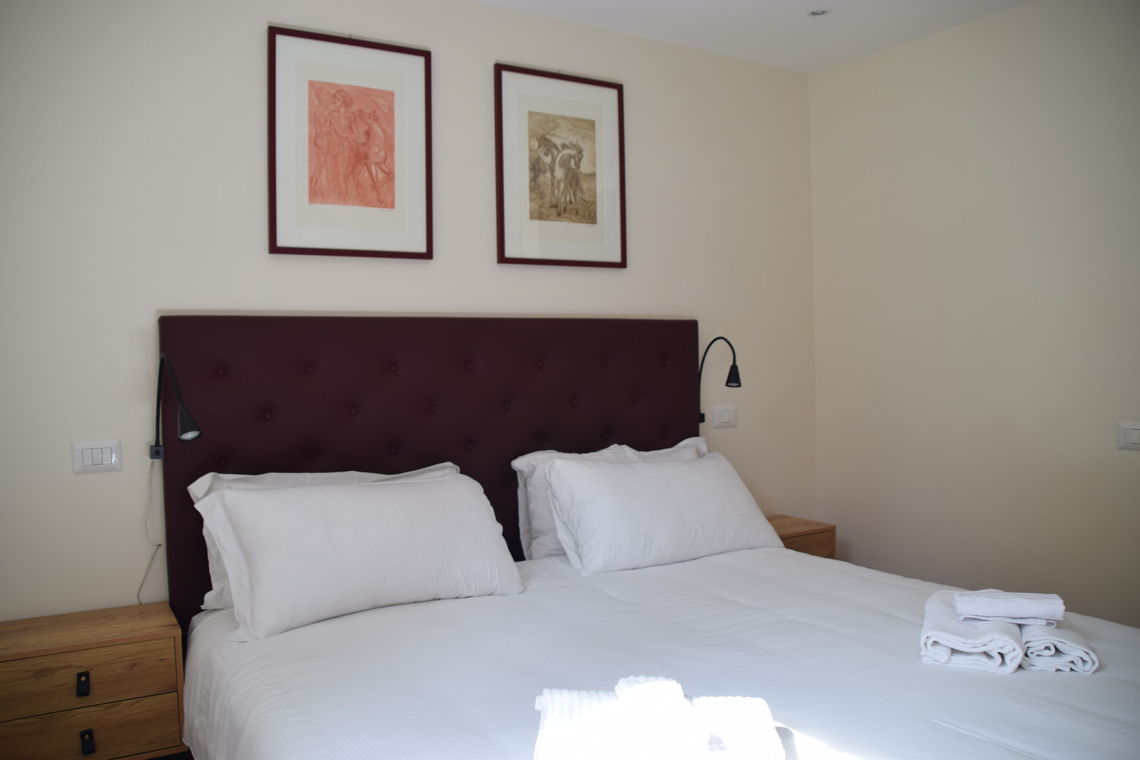 A neatly made bed with white linens and two pillows, a wooden nightstand with a lamp, and two framed artworks on the wall.