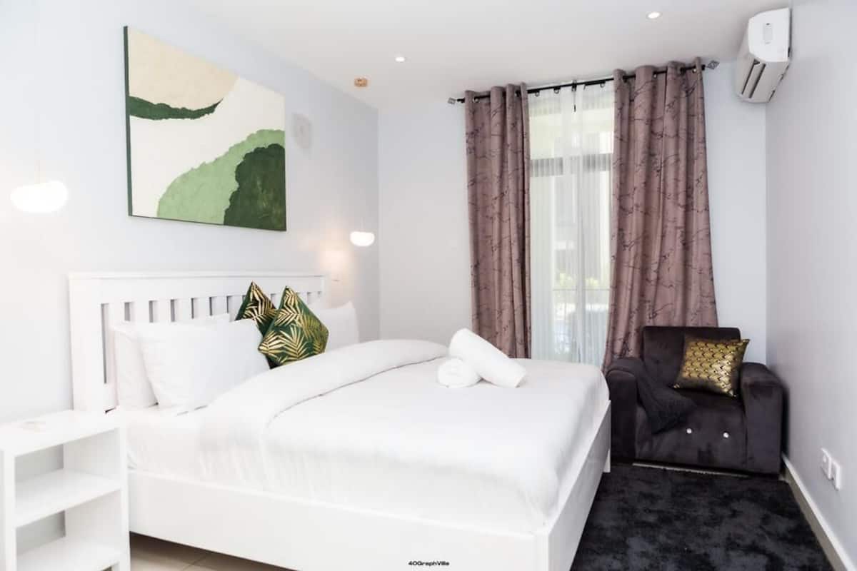 A modern hotel room with a neatly made bed, a small armchair, and a large window with curtains.