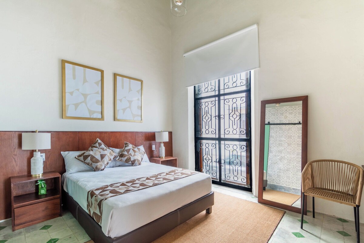 Un dormitorio con cama, mesitas de noche, una silla y un ventanal con un panel decorativo.