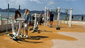 Ein Fitnessbereich im Freien mit Trainingsgeräten, einem befestigten Bereich und Blick auf das Meer und die Berge.