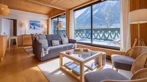Ett mysigt vardagsrum med en soffa, fåtöljer, ett soffbord i glas och utsikt över snöklädda berg genom stora fönster.