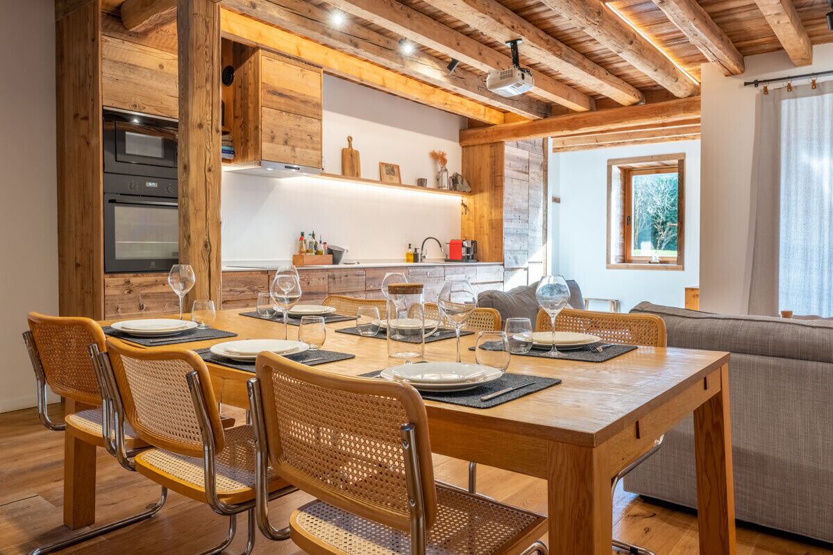 Une table à manger en bois, avec des chaises en osier, pouvant accueillir six personnes, une cuisine avec des meubles en bois et une fenêtre donnant sur un paysage arboré.