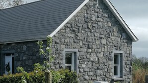 A stone house with a sloped roof, two windows, and a small garden area with a wooden post and a hedge.