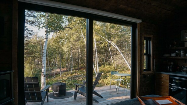 Una cabaña con un amplio ventanal corredera que da a una terraza exterior. En su interior, hay una cama, una pequeña mesa y una kitchenette con estufa.