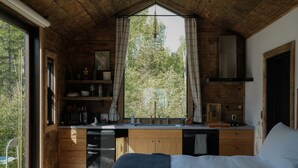 Un dormitorio compacto con techo de madera, una cocina integrada con fregadero y armarios, y un amplio ventanal con vistas a árboles.