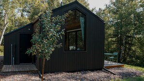A modern, dark-colored cabin with a large window and a wooden deck, surrounded by trees and gravel.