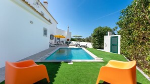 Ein Swimmingpool mit zwei orangefarbenen Stühlen, ein weißes Gebäude mit grüner Tür und klarer blauem Himmel.