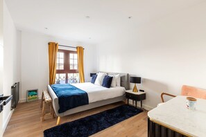 A modern bedroom with a bed, bedside table, lamp, chair, and a view of the city through a window. - City View Spacious 2 Bedroom Penthouse (London)