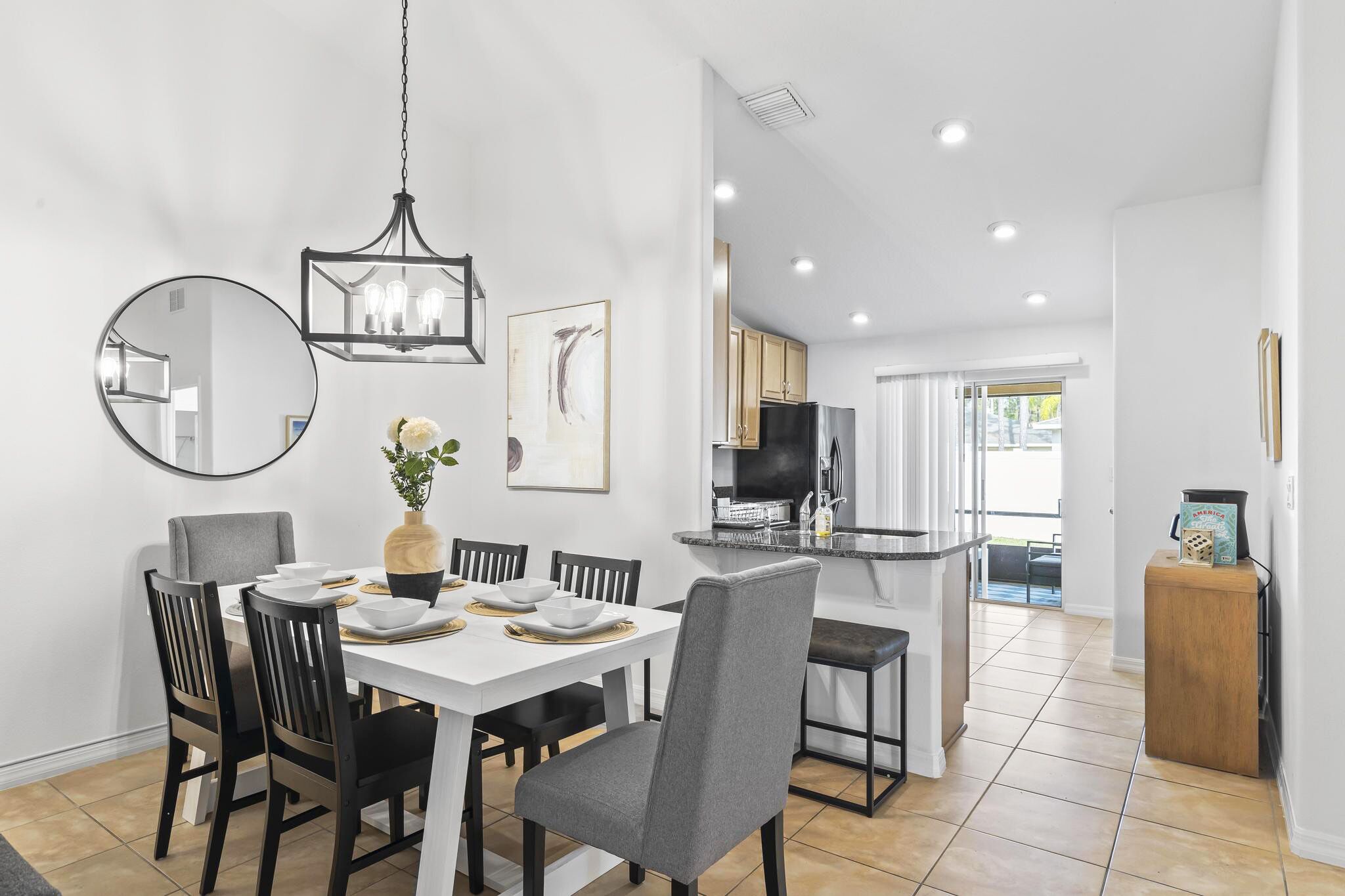 A modern dining area with a white table, black chairs, and a chandelier. There is a kitchen in the background with a refrigerator and a television.