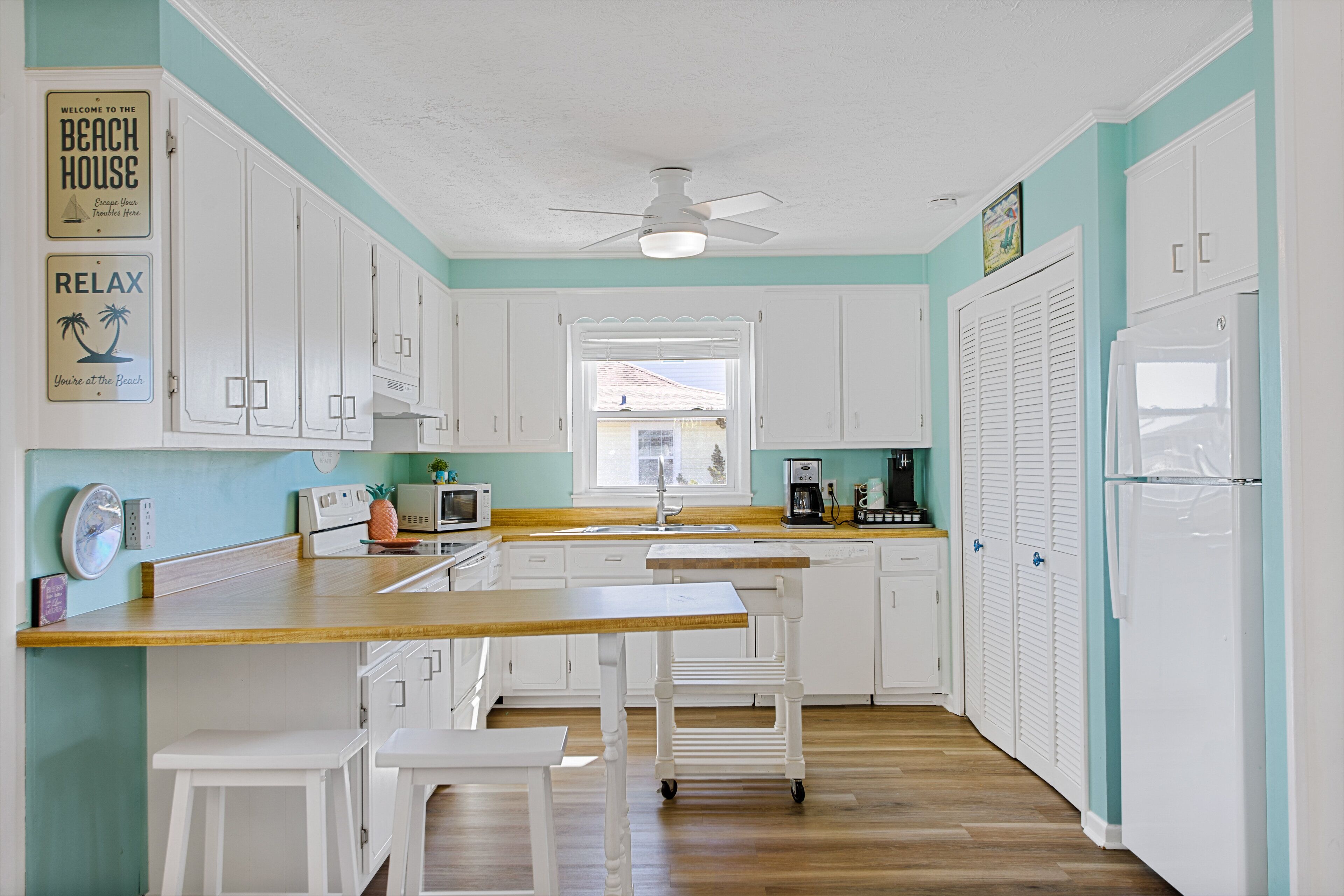A kitchen with white cabinets, a wooden countertop, a ceiling fan, and a window with a view of a house.