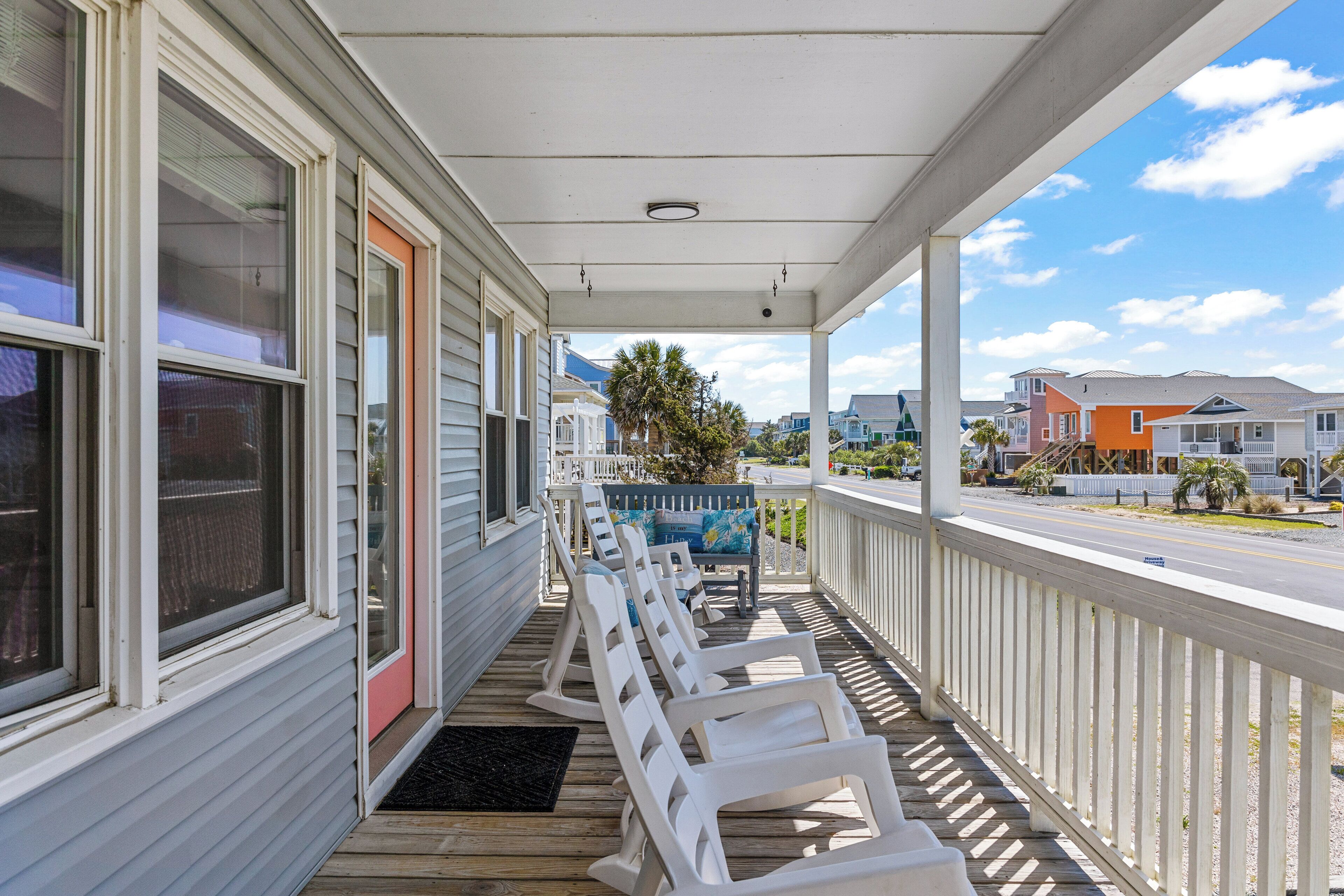 A covered porch with white chairs and a bench, overlooking a street with houses.