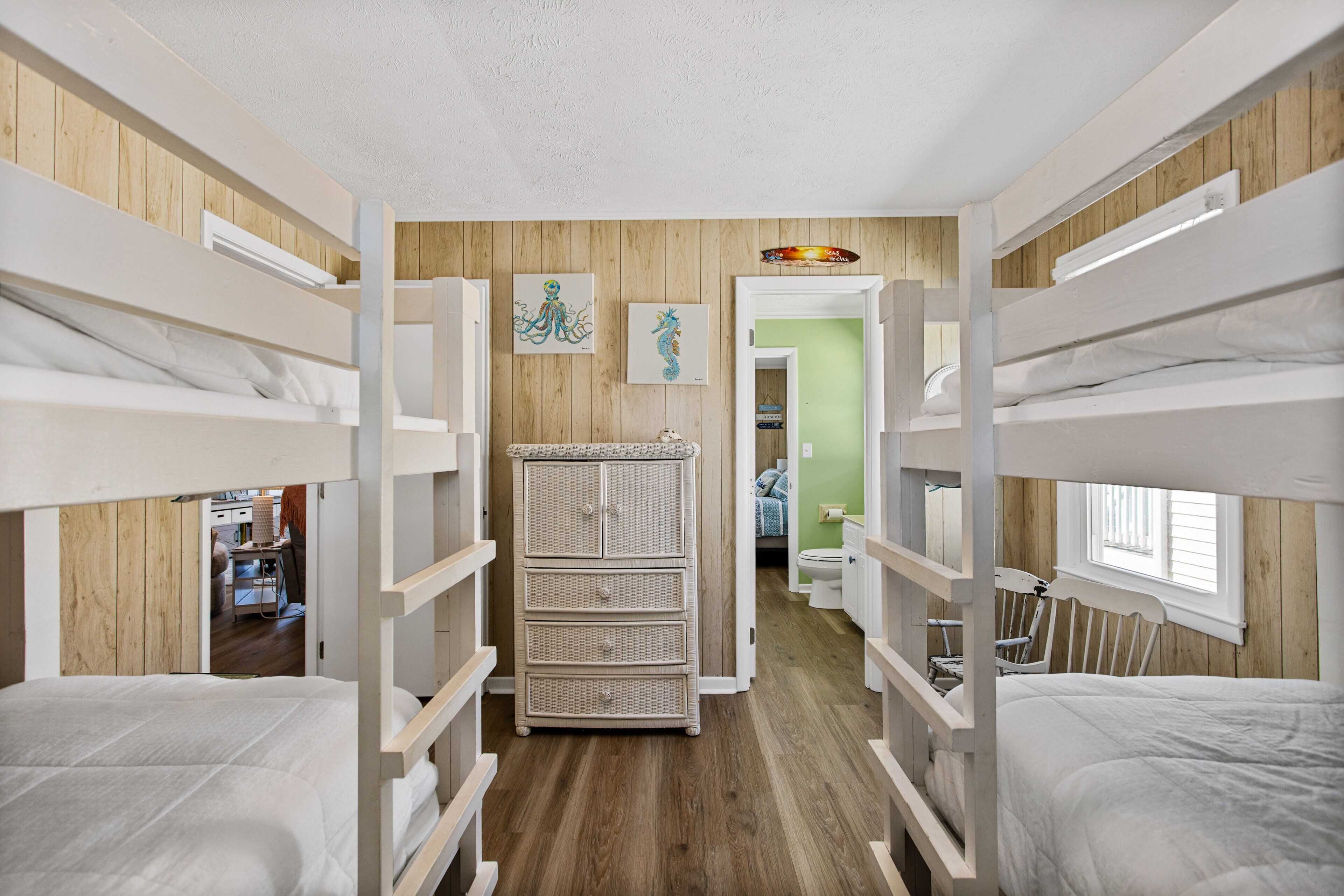 A bunk room with wooden walls, a dresser, and a view into another room.