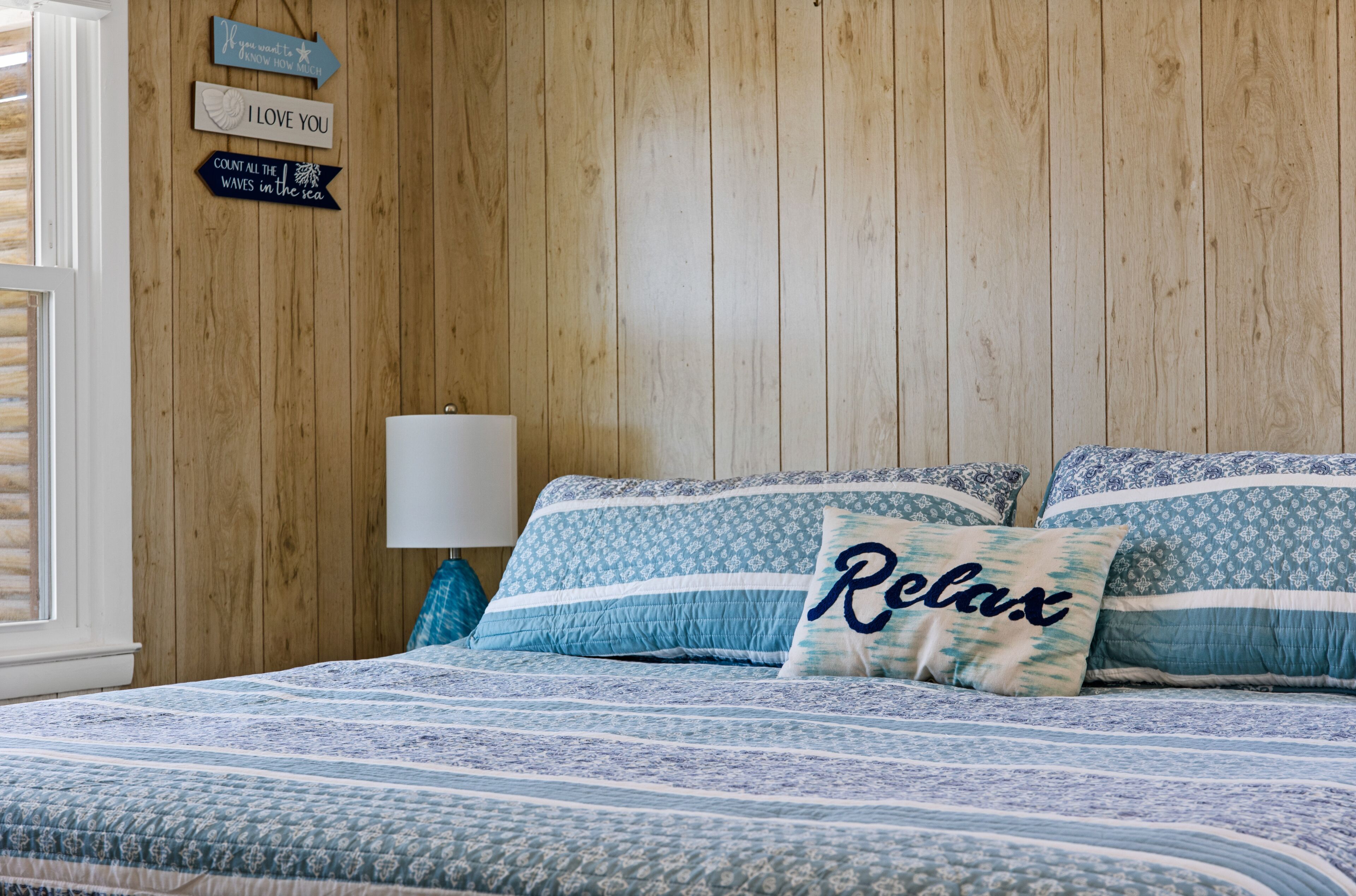 Een slaapkamer met een houten wand, een bed met blauw beddengoed en een kussen met de tekst 'Relax'.