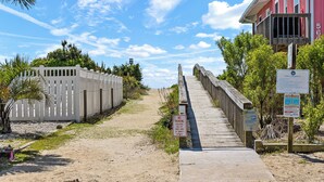 Eine Holzpromenade führt zu einem Strandabschnitt mit einem weißen Zaun und einem roten Gebäude im Hintergrund.