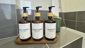 Tres botellas de gel de baño, acondicionador y champú sobre una bandeja de madera en un baño con paredes de azulejos y ducha.