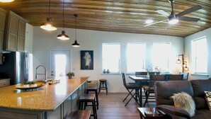 A modern kitchen with a central island, wooden ceiling, and pendant lighting. There is a dining area with a table and chairs, and a living room with a sofa and a fireplace.