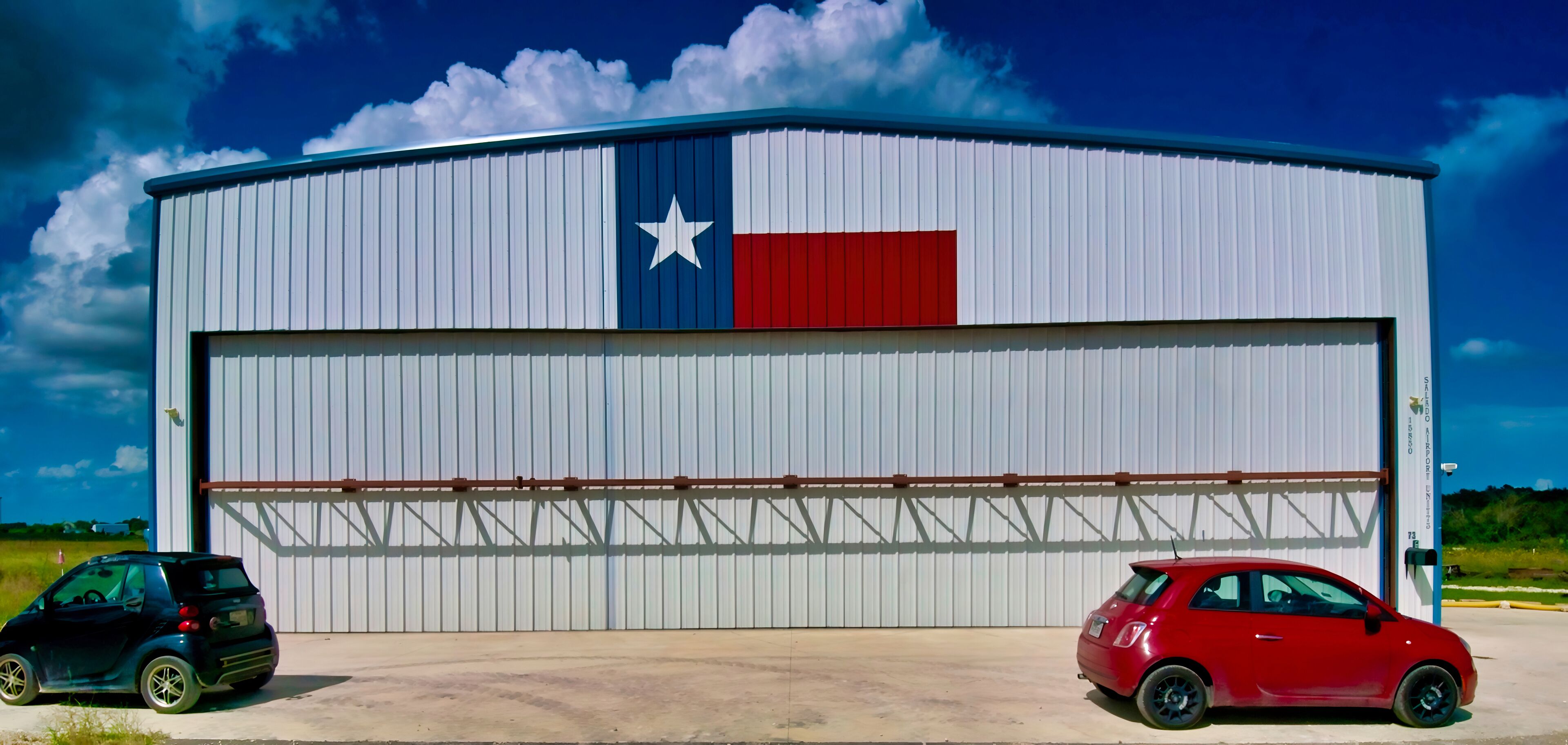 A large industrial building with a Texas flag design, featuring two cars parked in front.