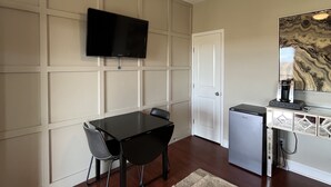 A hotel room with a dining table, chairs, a TV mounted on the wall, a ceiling fan, a refrigerator, and a patterned rug. - Lonely Oak Guesthouse! Room 4 (Llano County)