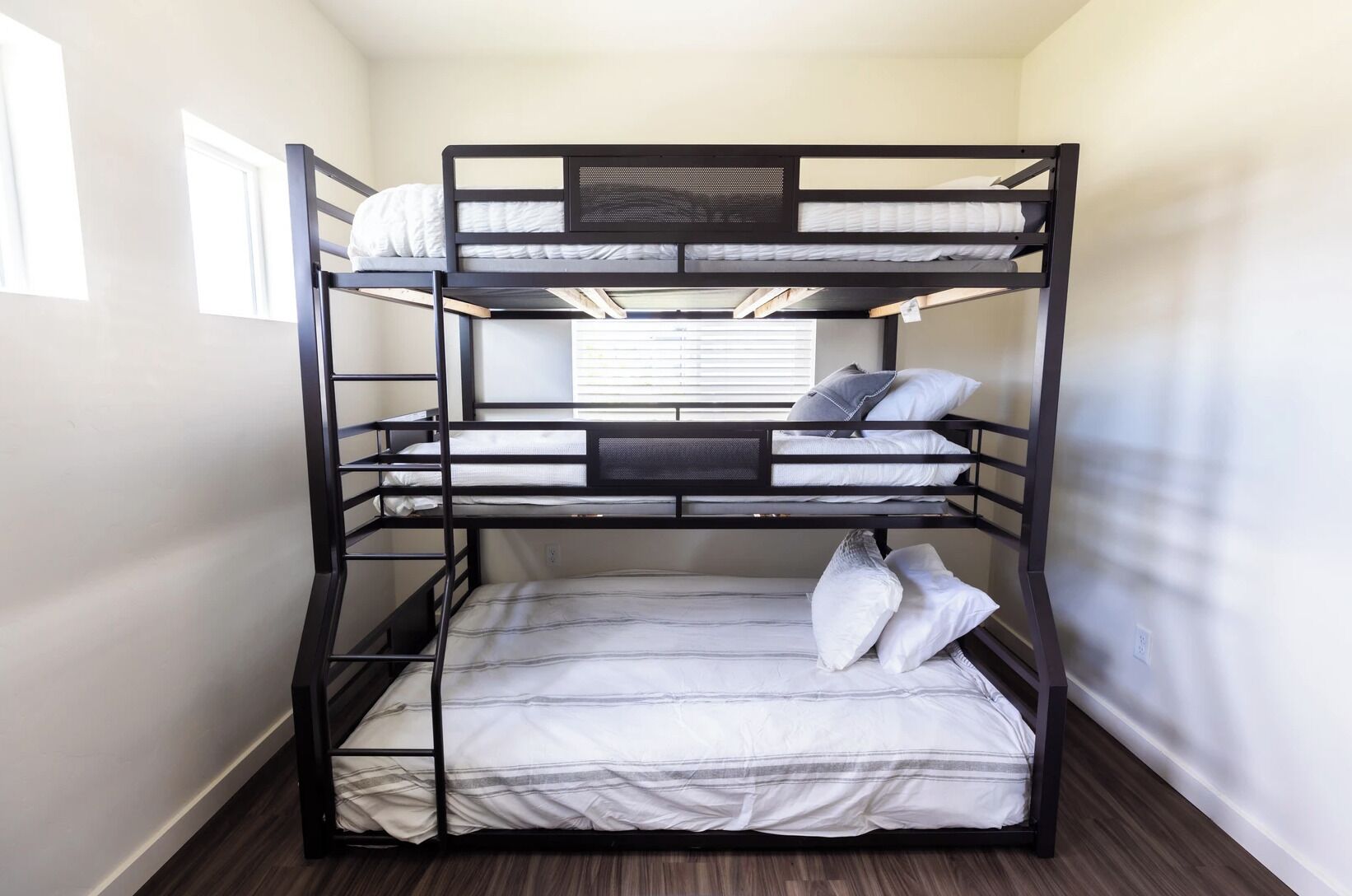 Een stapelbed met drie niveaus, een raam met zonwering en een houten vloer.