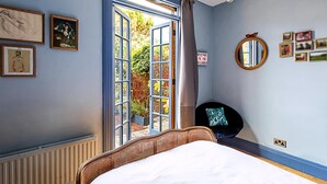 A bedroom with a large window, a bed, a chair, and wall decorations including framed pictures and a mirror. - Charming Acton Dollhouse (London)