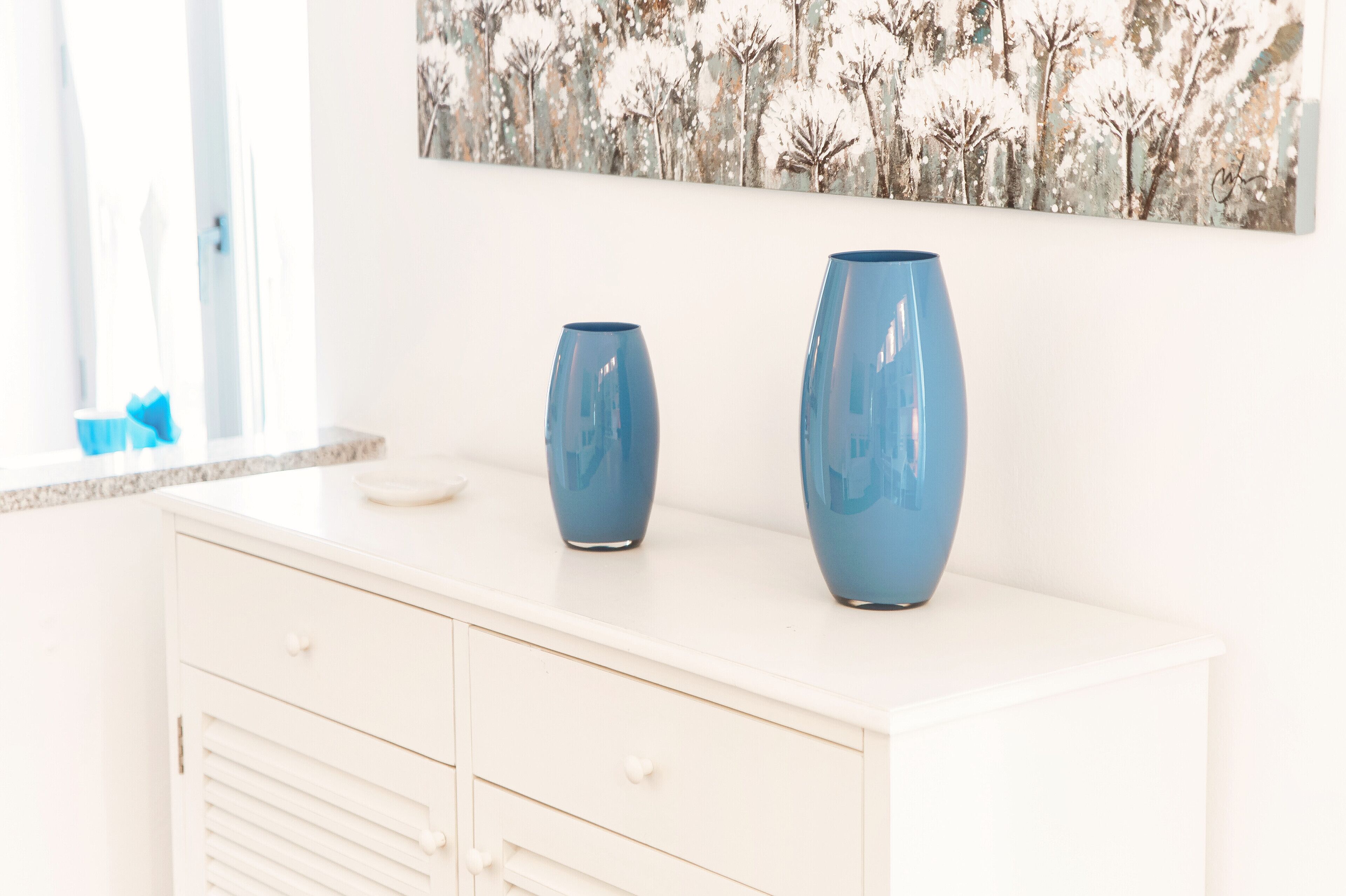 A white cabinet with two blue vases on top and a painting of trees on the wall behind.