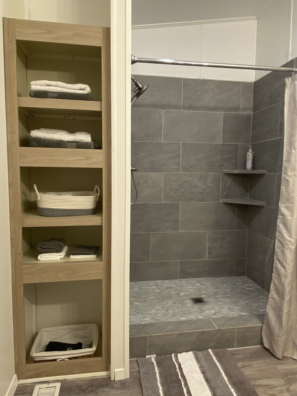 Een inloopdouche met grijze tegels, een houten plank met handdoeken, en een gestreept vloerkleed.