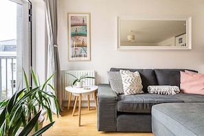 A modern living room with a grey sofa, a small white table, a potted plant, and a framed artwork on the wall. - Bright Flat in Historic Whitechapel (London)
