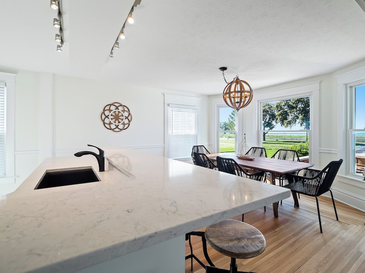 A modern kitchen with a large island, a dining area with a wooden table and chairs, and large windows with a view of greenery.