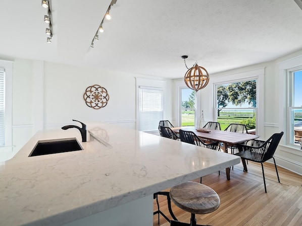 A modern kitchen with a large island, a dining area with a wooden table and chairs, and large windows with a view of greenery.