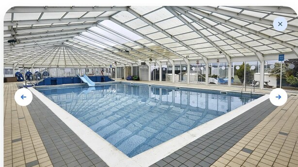 A large indoor swimming pool with a glass ceiling, surrounded by tiled flooring and a blue slide.