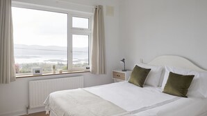 Un dormitorio con un ventanal grande, una cama con ropa de cama blanca y una mesita de noche con lámpara.
