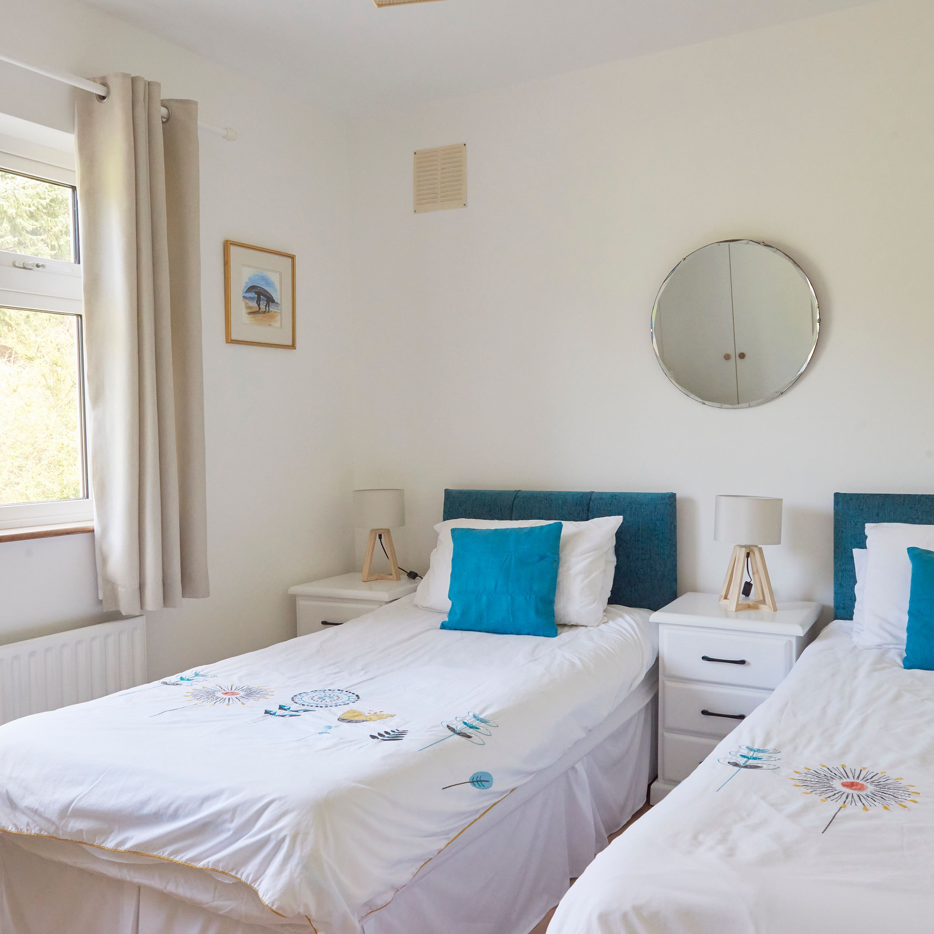 A bedroom with two beds, a round mirror, and a window with curtains.